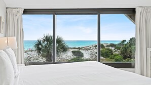 Un dormitorio con una cama grande, un ventilador de techo y vistas a una playa y al océano a través de ventanales de pared a pared.