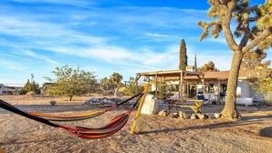 A desert landscape with a hammock, a covered outdoor seating area, and a building with a white exterior. - Dez-Art Retreat - Eclectic Getaway near JTNP (Yucca Valley)