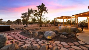 An outdoor patio area with stone pathways, seating, and a pergola with string lights. - Dez-Art Retreat - Eclectic Getaway near JTNP (Yucca Valley)