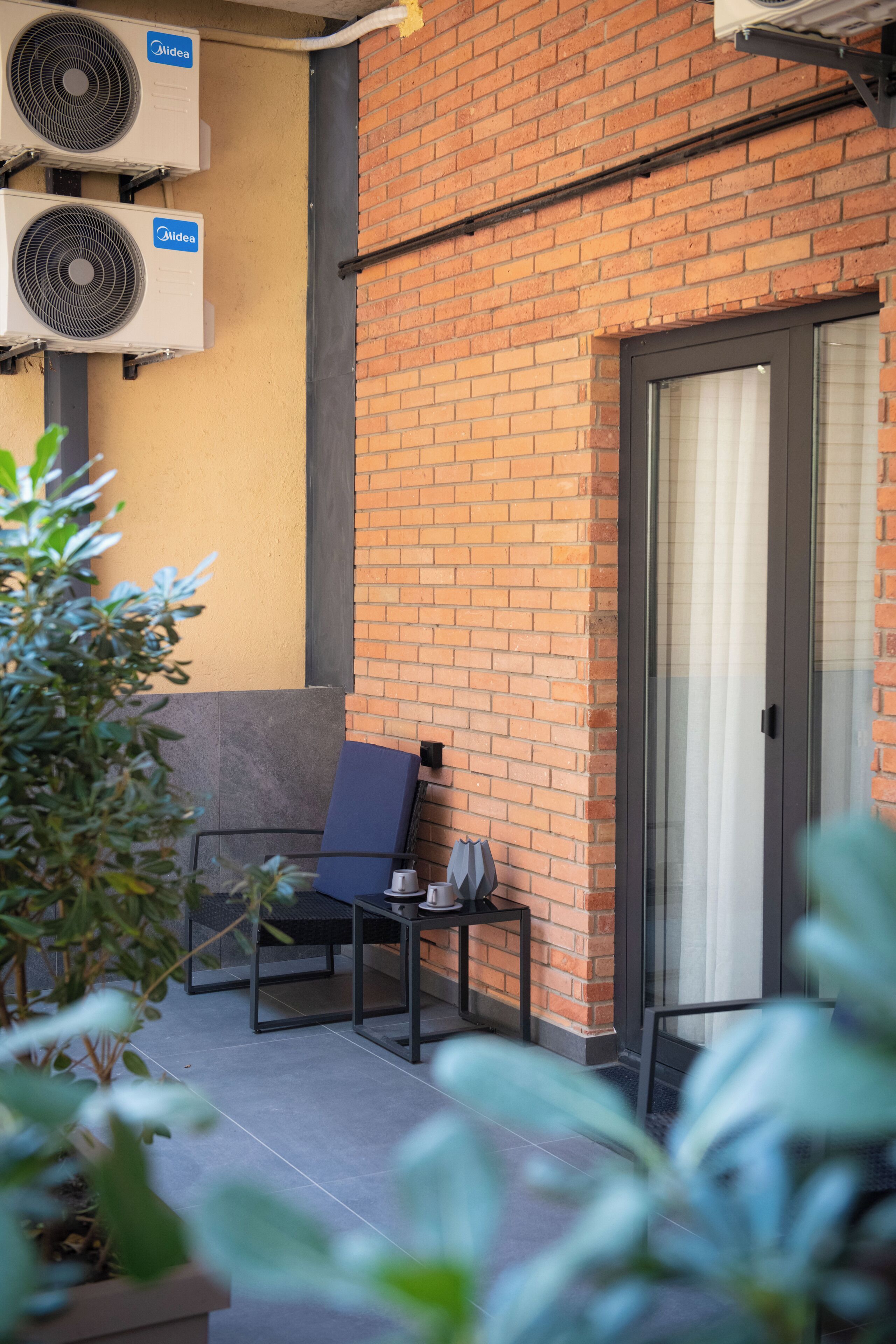 An outdoor seating area with a brick wall, two air conditioning units, a door, and potted plants.