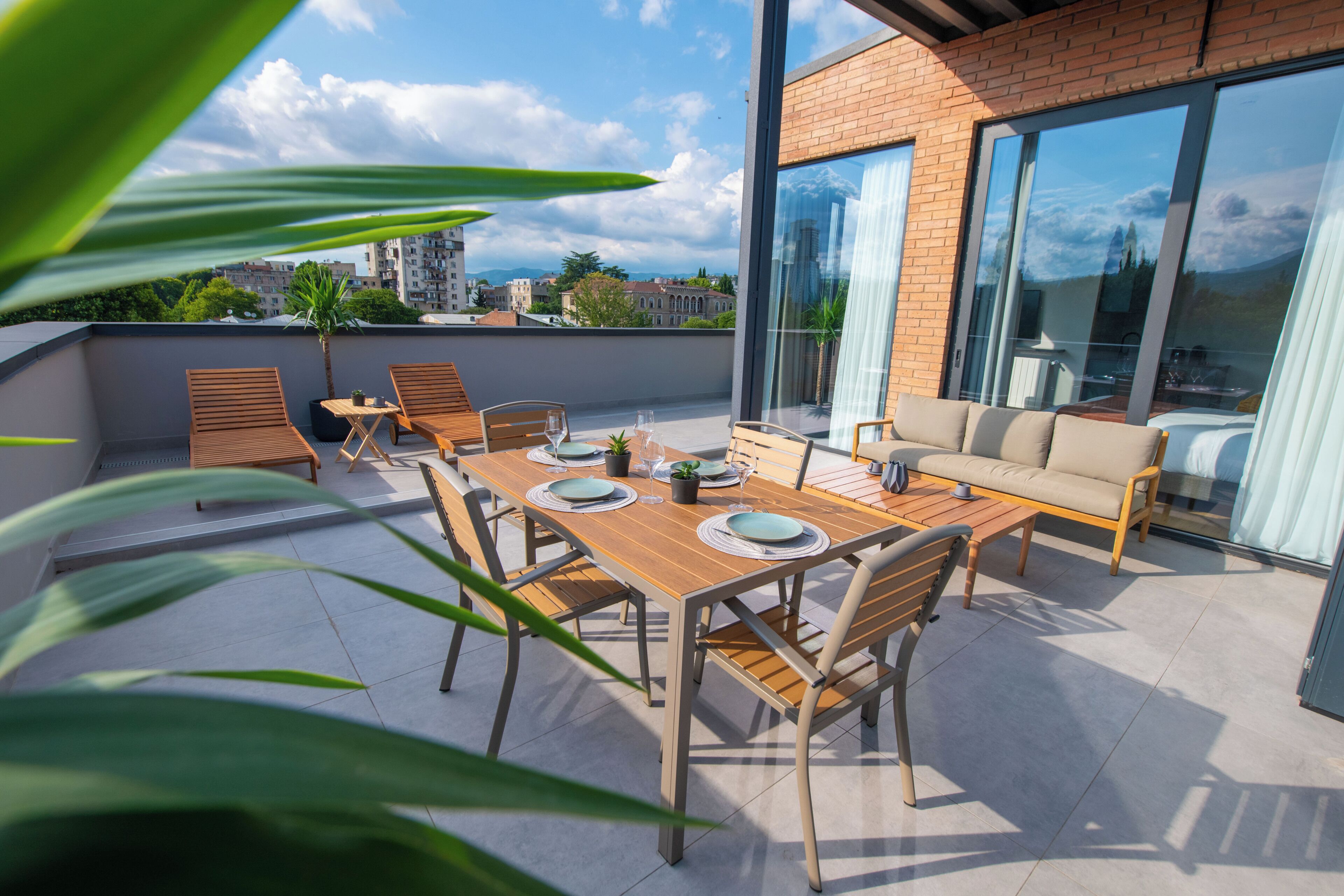 Una terraza en la azotea con una mesa de madera dispuesta para comer, un sofá y una pared de vidrio que ofrece vistas a la ciudad.