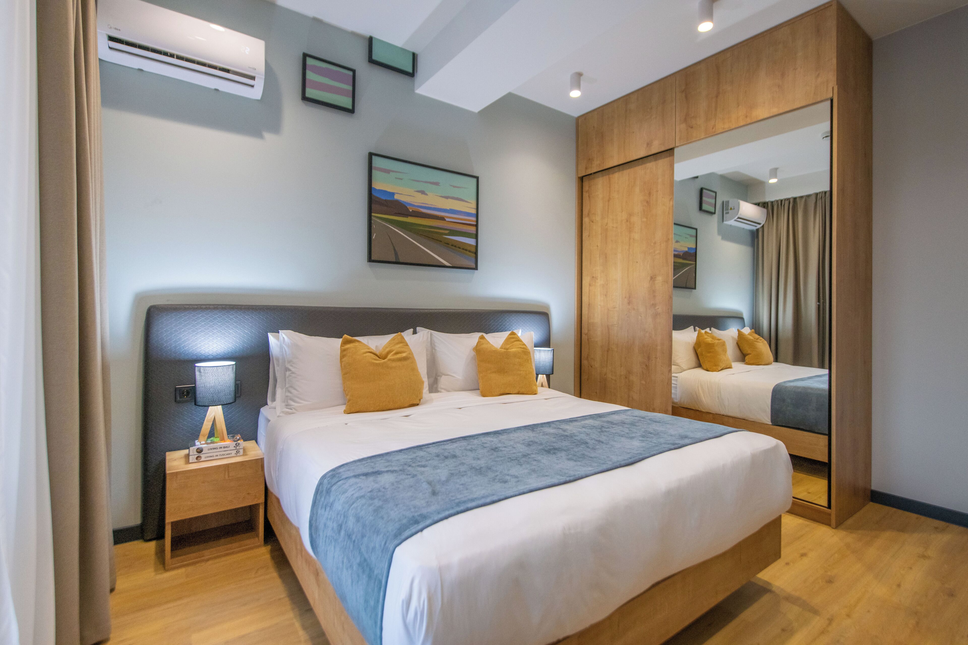 A hotel room with a large bed, a nightstand with a lamp, an air conditioning unit, a painting on the wall, and a mirror.