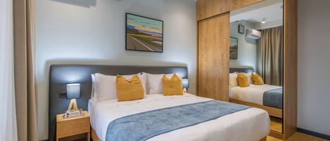 A hotel room with a large bed, a nightstand with a lamp, an air conditioning unit, a painting on the wall, and a mirror.