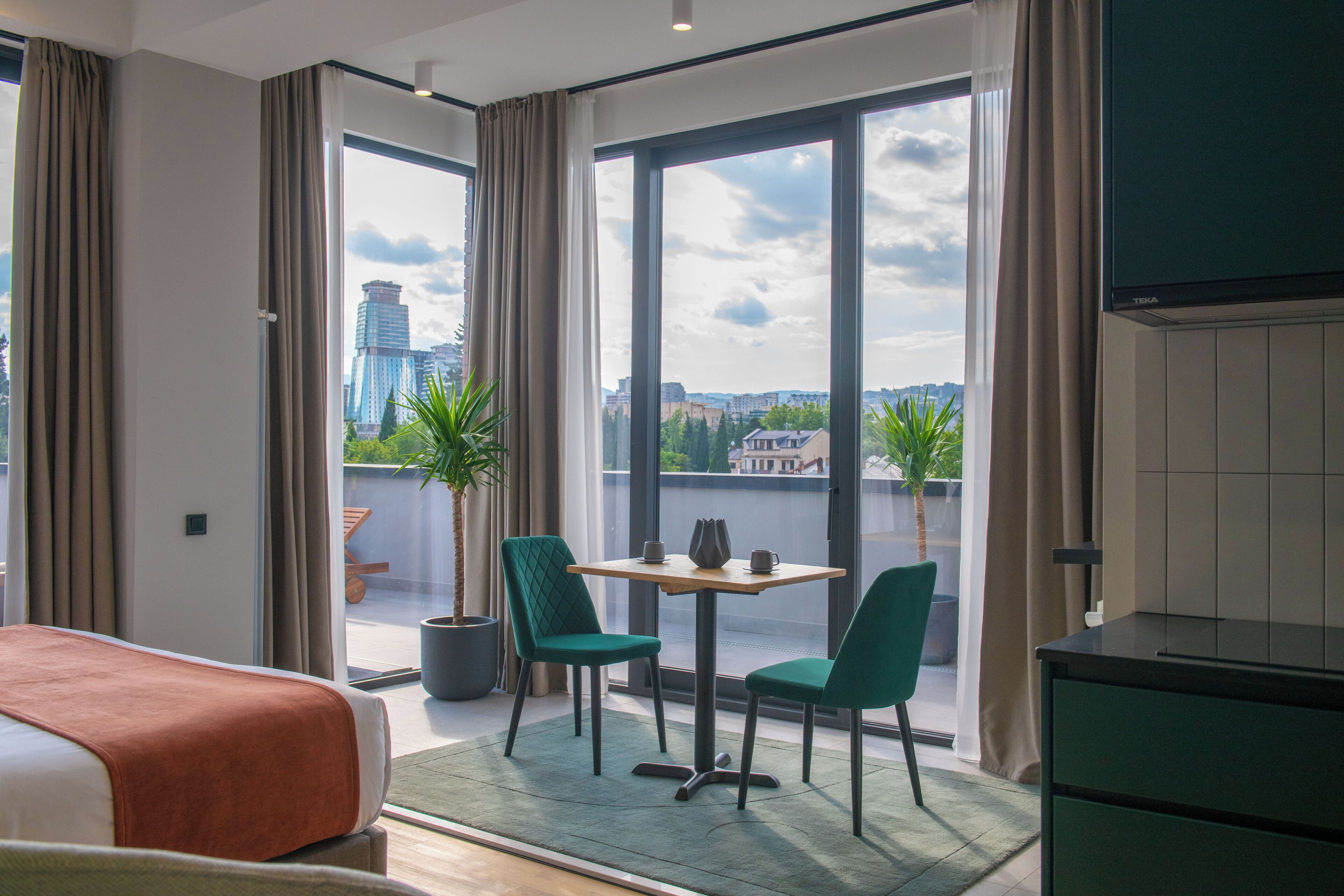 Une chambre d’hôtel comprenant un lit, une table à manger avec des chaises, un téléviseur et une vue sur le paysage urbain grâce à de grandes fenêtres.