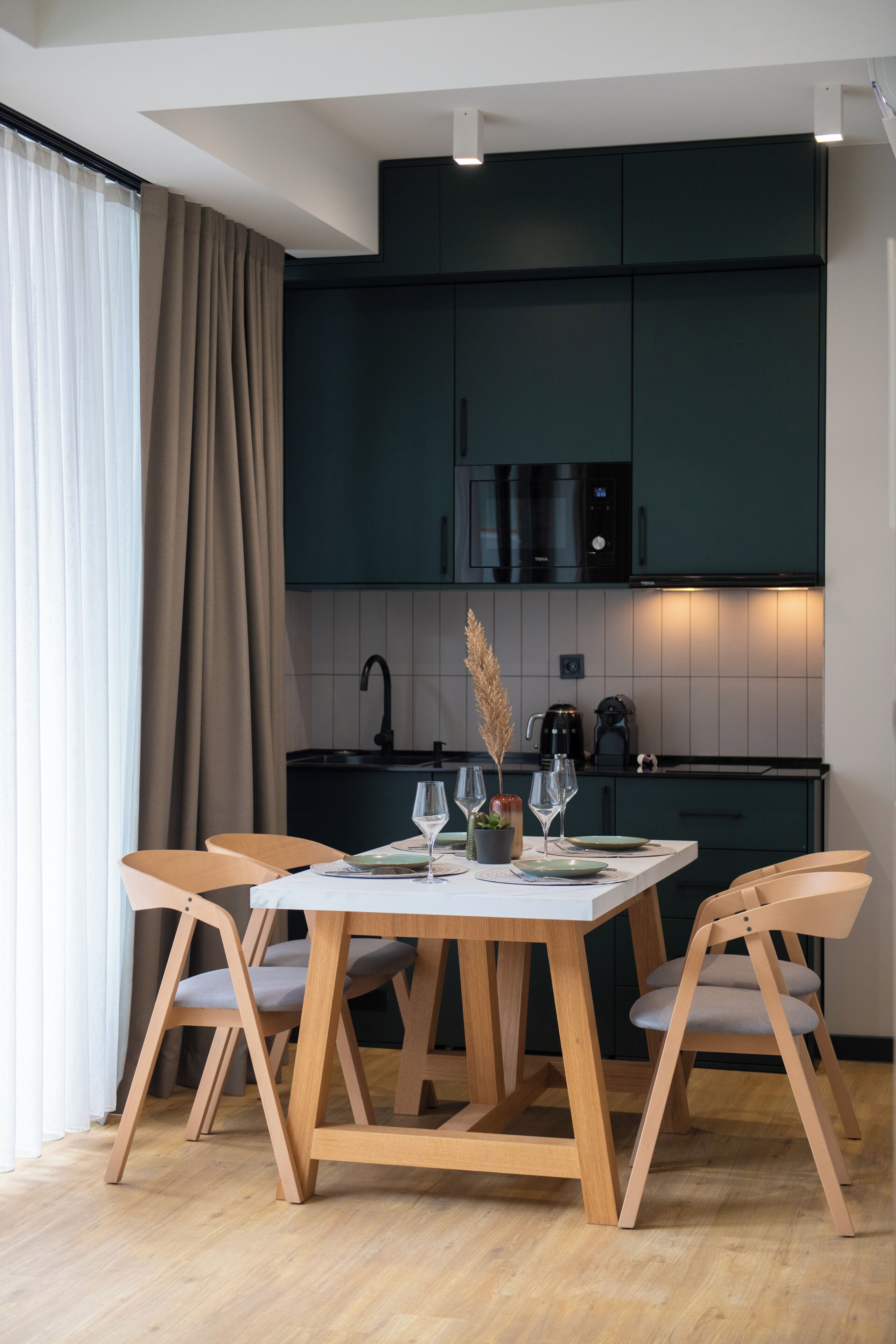 Una cocina moderna con armarios oscuros, una encimera blanca y una zona de comedor con sillas de madera y una mesa preparada para dos.