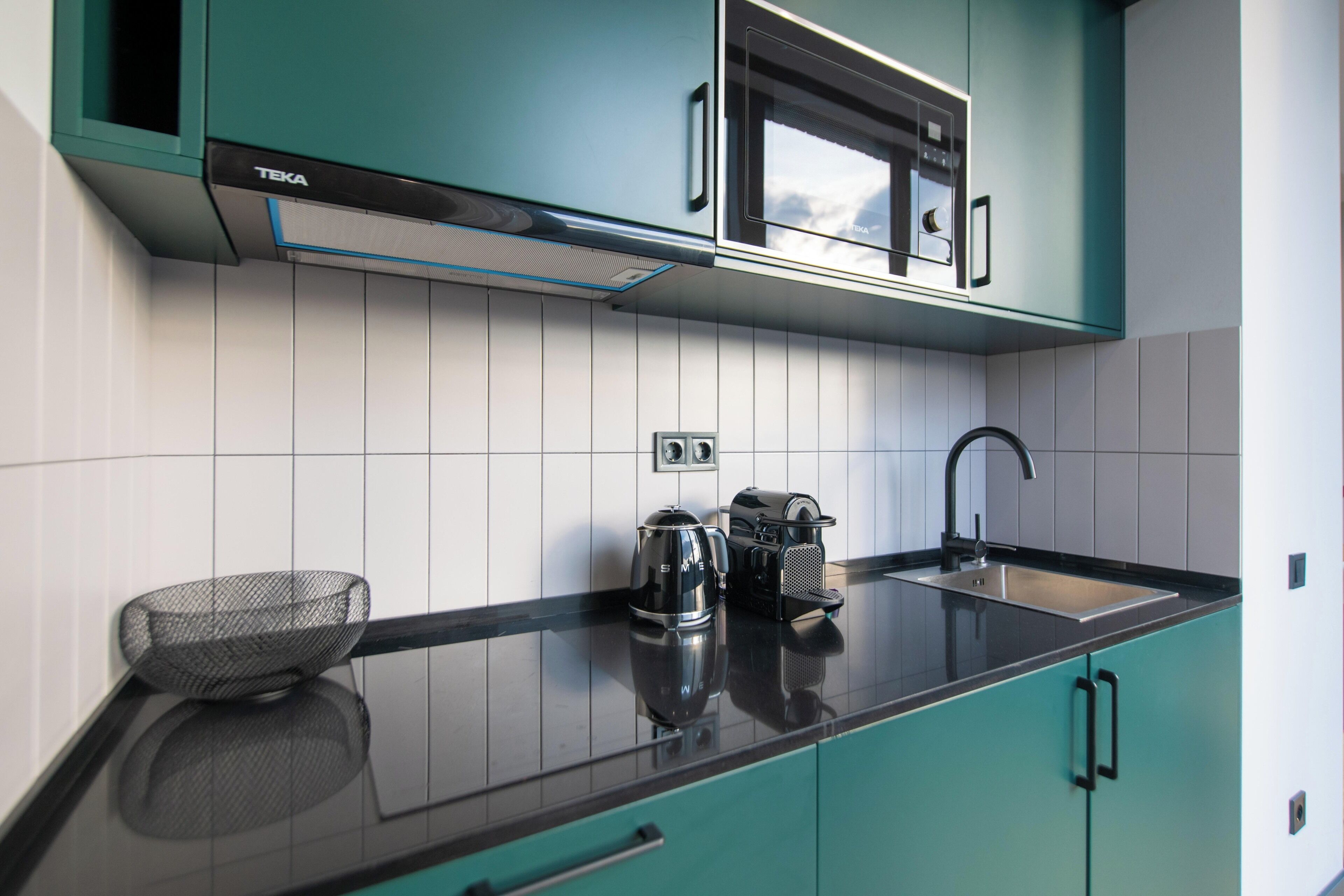 A modern kitchen with teal cabinets, a black countertop, a coffee maker, and a kettle.