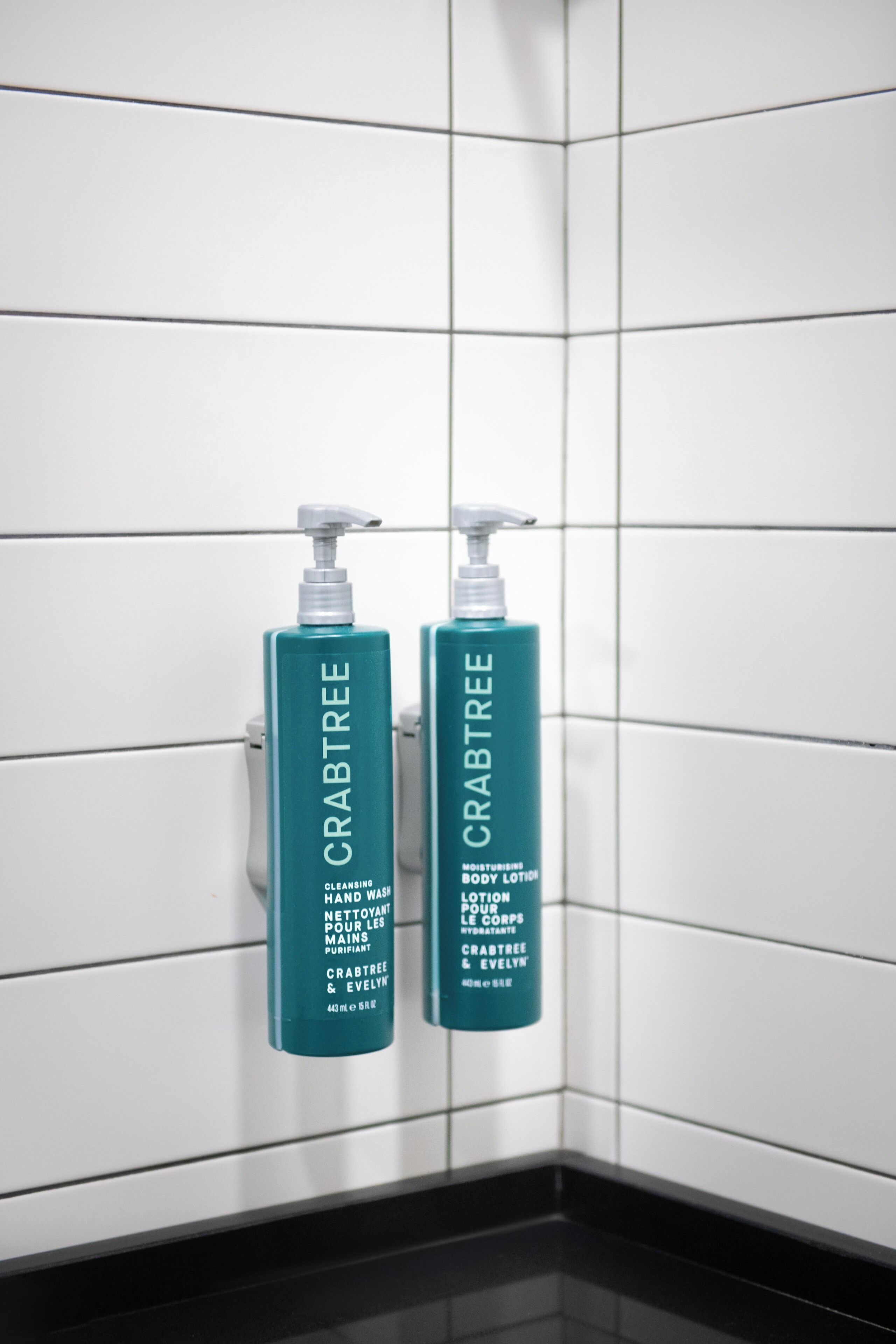 Two bottles of Crabtree body lotion on a white tiled wall.