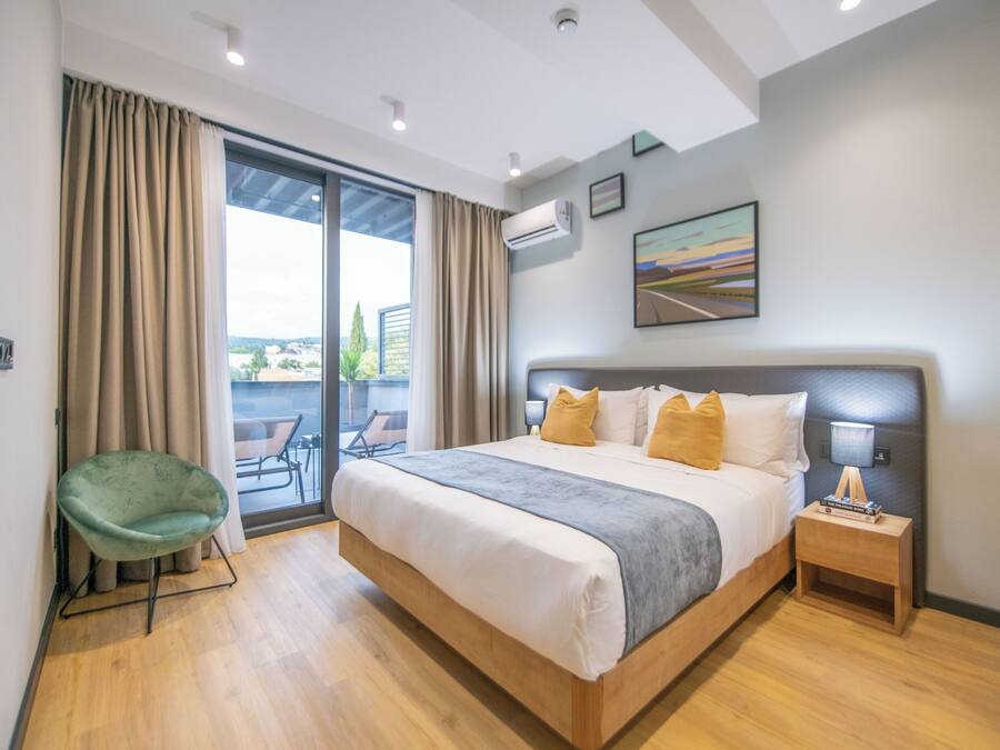 A modern hotel room with a large bed, a green chair, a small bedside table, and a view of the outdoors through a sliding glass door.