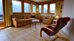 A cozy living room with a sofa, a rocking chair, a small table, and large windows offering a mountain view. - Exterior : Apartment on the lower ground floor of a building, for 4 people close to the hotel/restau (St-Luc)