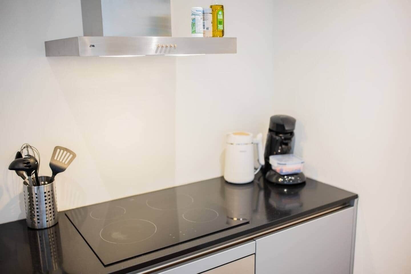 A kitchen with a black countertop, a coffee maker, a white electric kettle, and a stainless steel range hood.