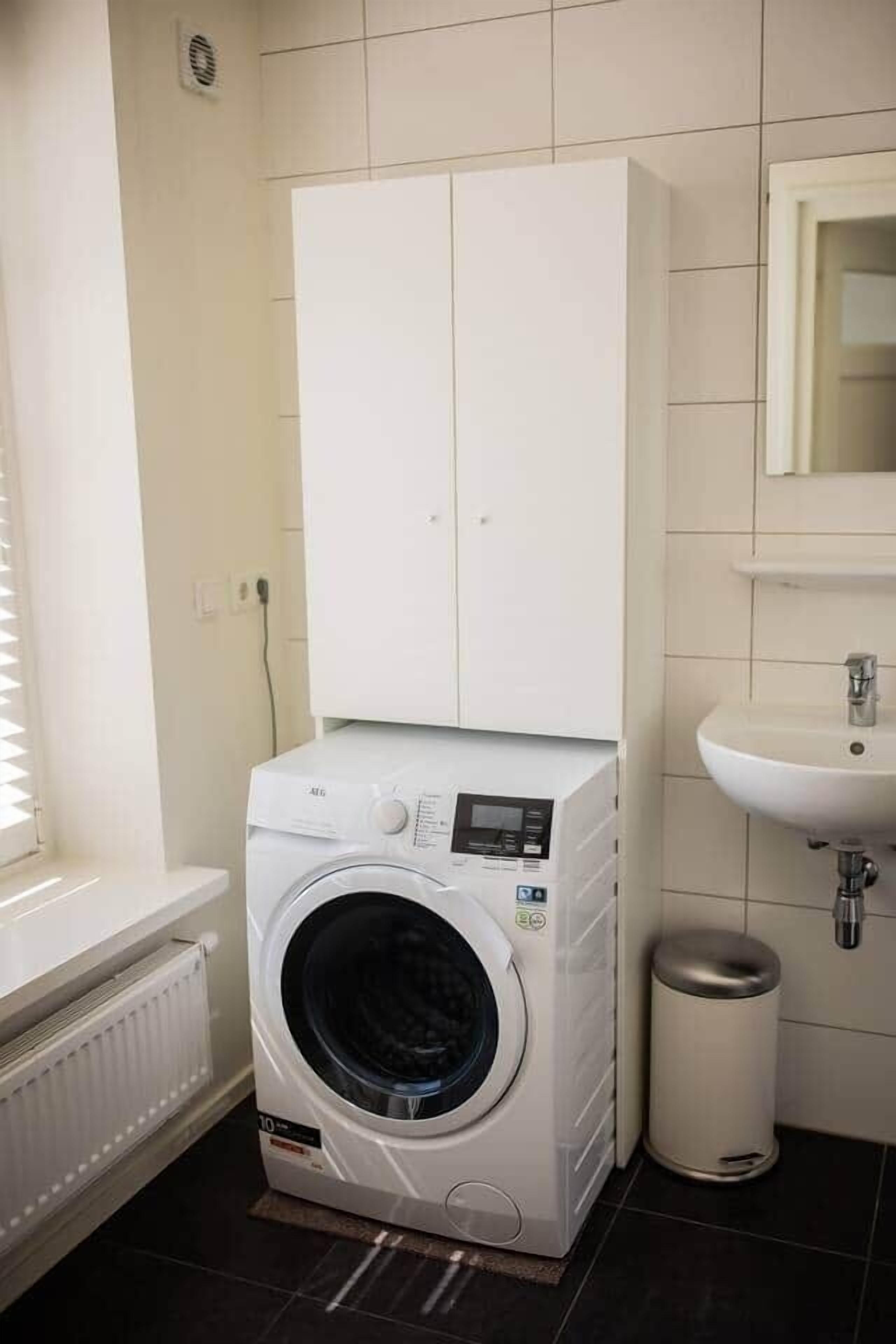 A compact laundry room with a front-loading washing machine, a small sink, and a trash bin.