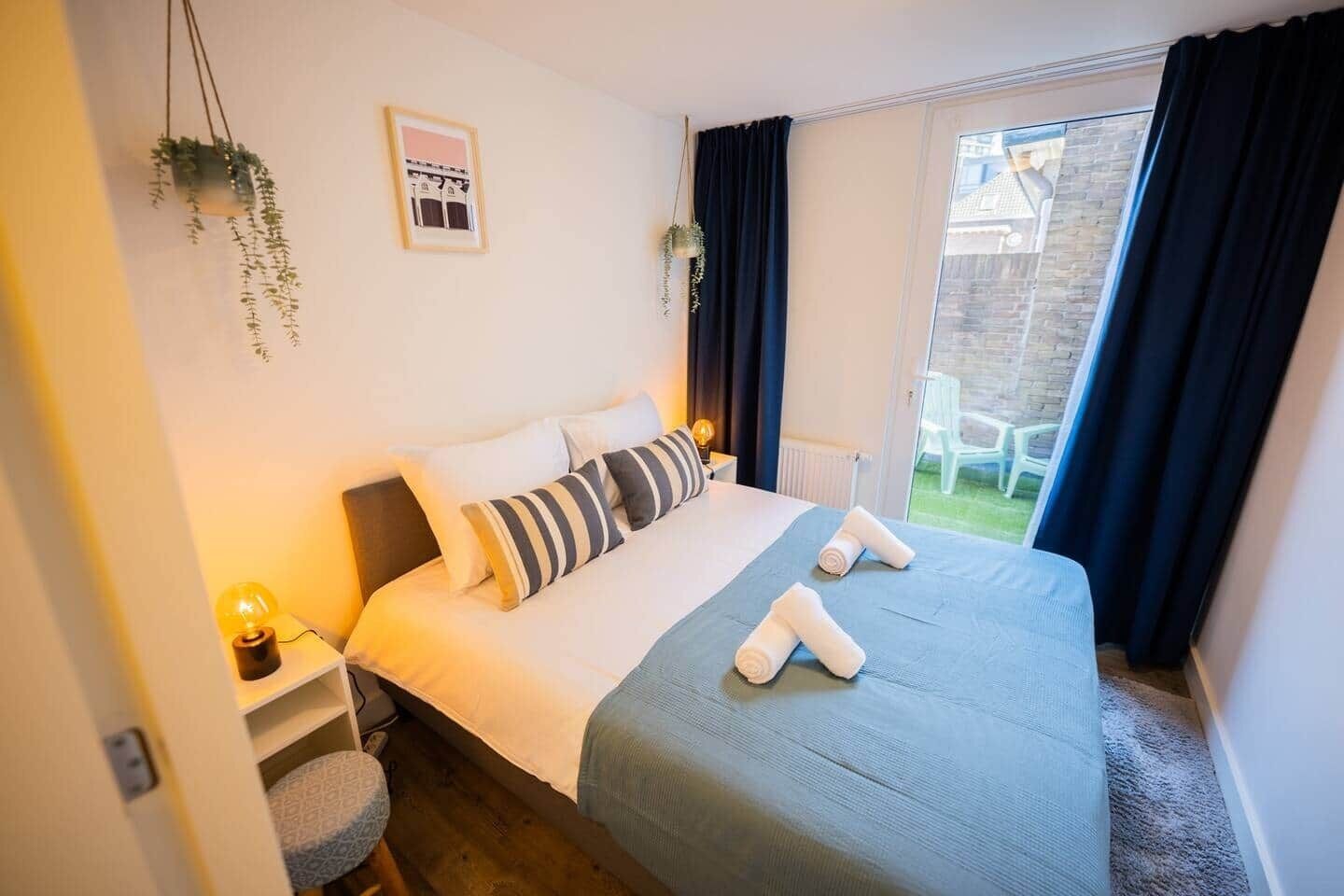 Un dormitorio con cama, almohadas, mesita de noche con lámpara, ventana con cortinas y una planta pequeña.