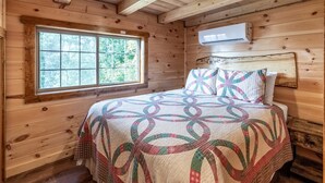 Ein Schlafzimmer einer Holzhütte mit einem großen Fenster, einem Bett mit einem gemusterten Quilz und einer Klimaanlage an der Wand.