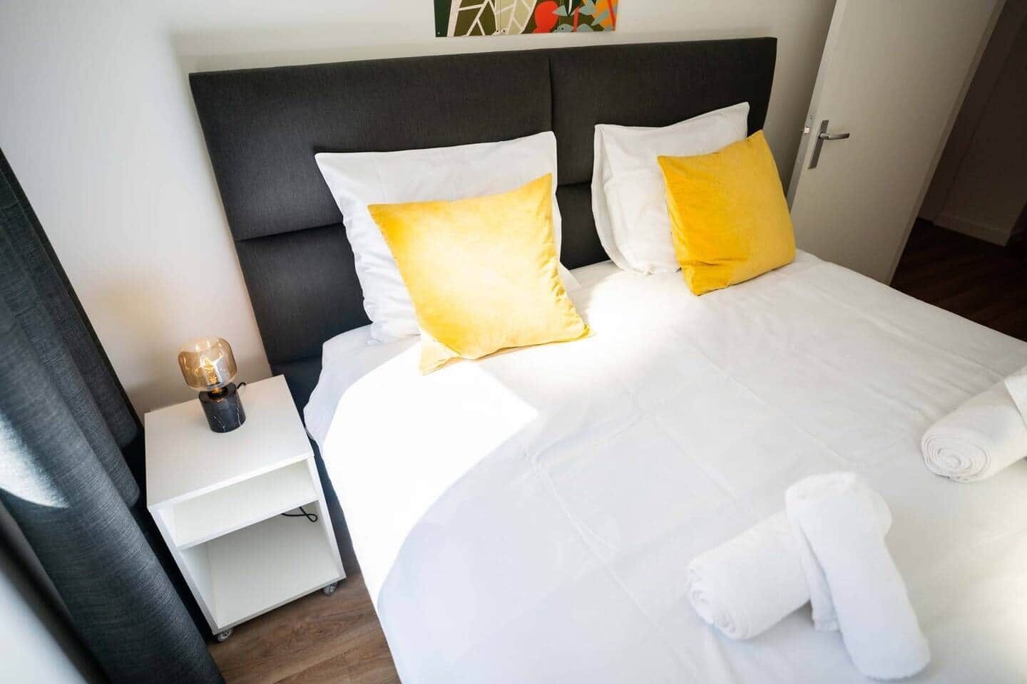 Et hotelværelse med en seng, hvide sengetøj, to gule puder, et natbord med en lampe og et sammenrullet håndklæde på sengen.