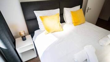 Een hotelkamer met een bed, wit beddengoed, twee gele kussens, een nachtkastje met een lamp, en een opgerolde handdoek op het bed.