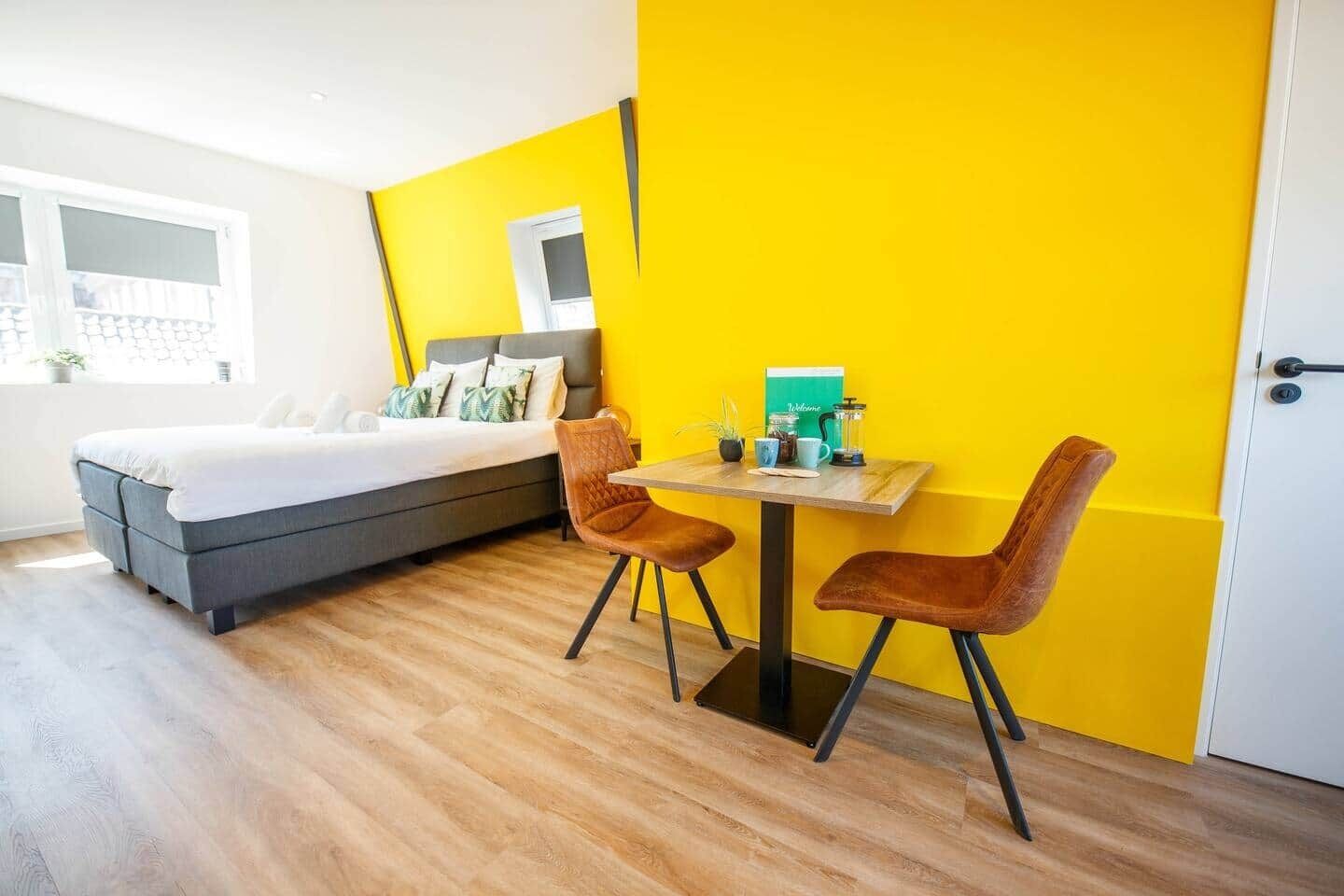 A modern room with a bed, two chairs, a small table with a coffee maker, and a bright yellow accent wall.