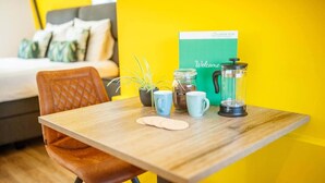 A wooden table with a coffee maker, two mugs, a jar of coffee, and a spoon in a room with a yellow wall and a bed with pillows in the background.