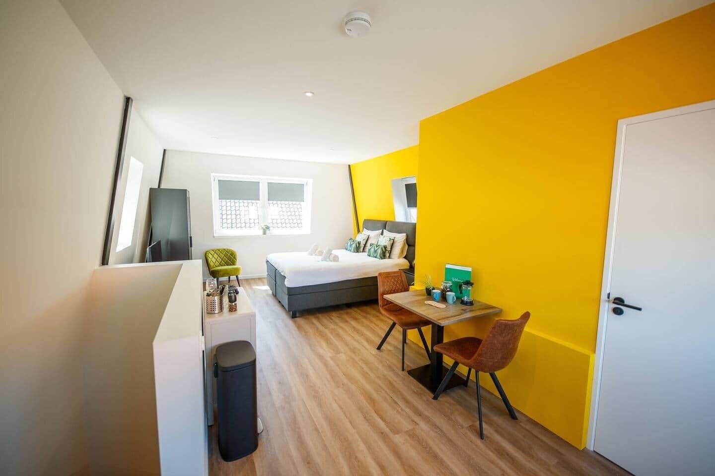 A compact hotel room with a bed, a small table, a chair, a desk, a trash bin, and a door.