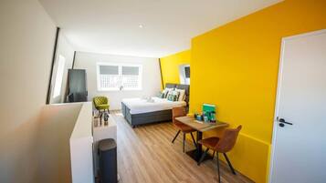 A compact hotel room with a bed, a small table, a chair, a desk, a trash bin, and a door.