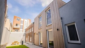 A modern urban courtyard with a wooden bench, a small grassy area, and a building with large windows and a grey facade. - Great 95m2 Two-Bedroom Apartment (WE-39) (Tiel)