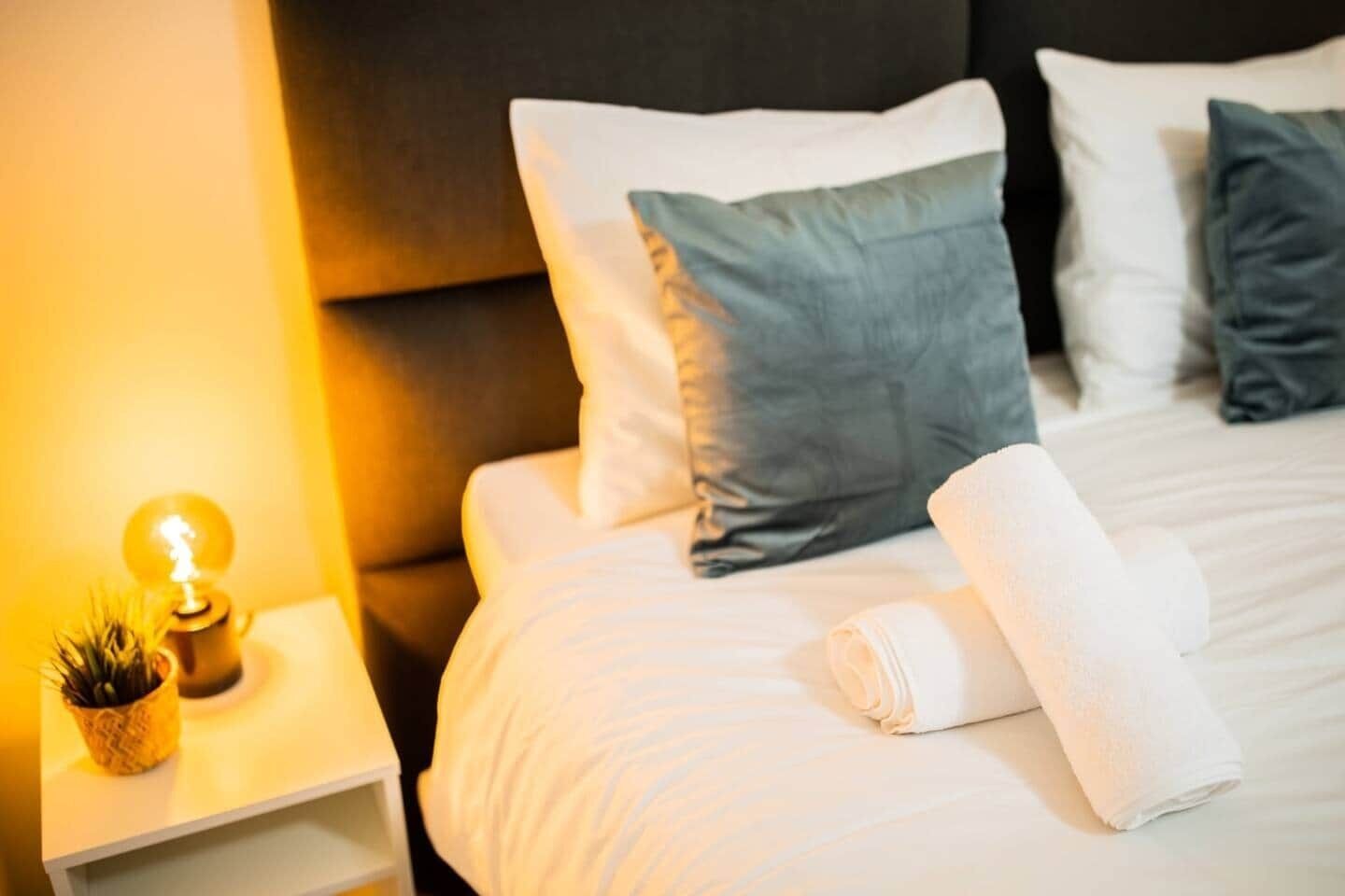 Una cama bien hecha con almohadas blancas y grises, una toalla blanca enrollada y una lámpara de noche con una pequeña planta.