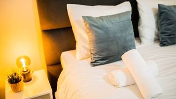 Una cama bien hecha con almohadas blancas y grises, una toalla blanca enrollada y una lámpara de noche con una pequeña planta.