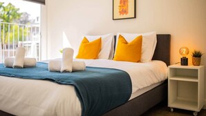 Una cama bien hecha con una manta azul y dos toallas enrolladas, una mesita de noche con una lámpara y una planta, y un cuadro en la pared.