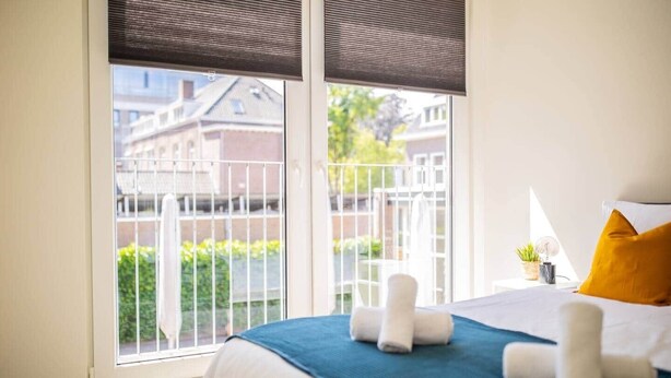 Un dormitorio con un ventanal grande, una cama con ropa de cama blanca y azul, un cojín amarillo y una mesita de noche con una lámpara y una planta.