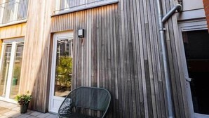 Un edificio moderno con fachada de madera, una puerta visible y un pequeño espacio al aire libre con mesas y sillas.