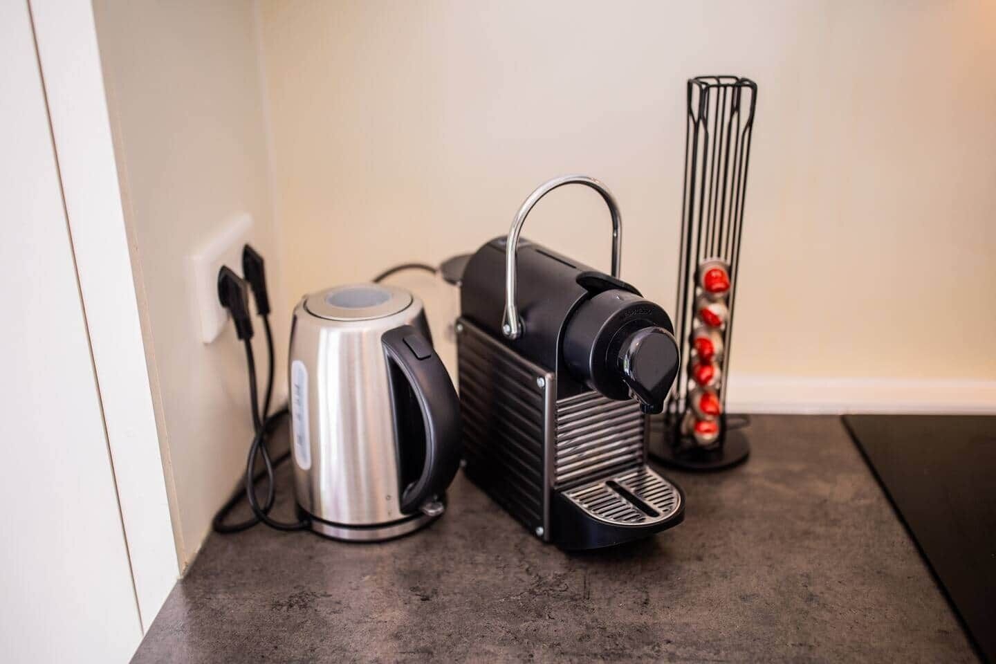 A kitchen with a kettle, a coffee maker, and a glass container with red objects.
