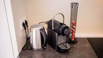 A kitchen with a kettle, a coffee maker, and a glass container with red objects.