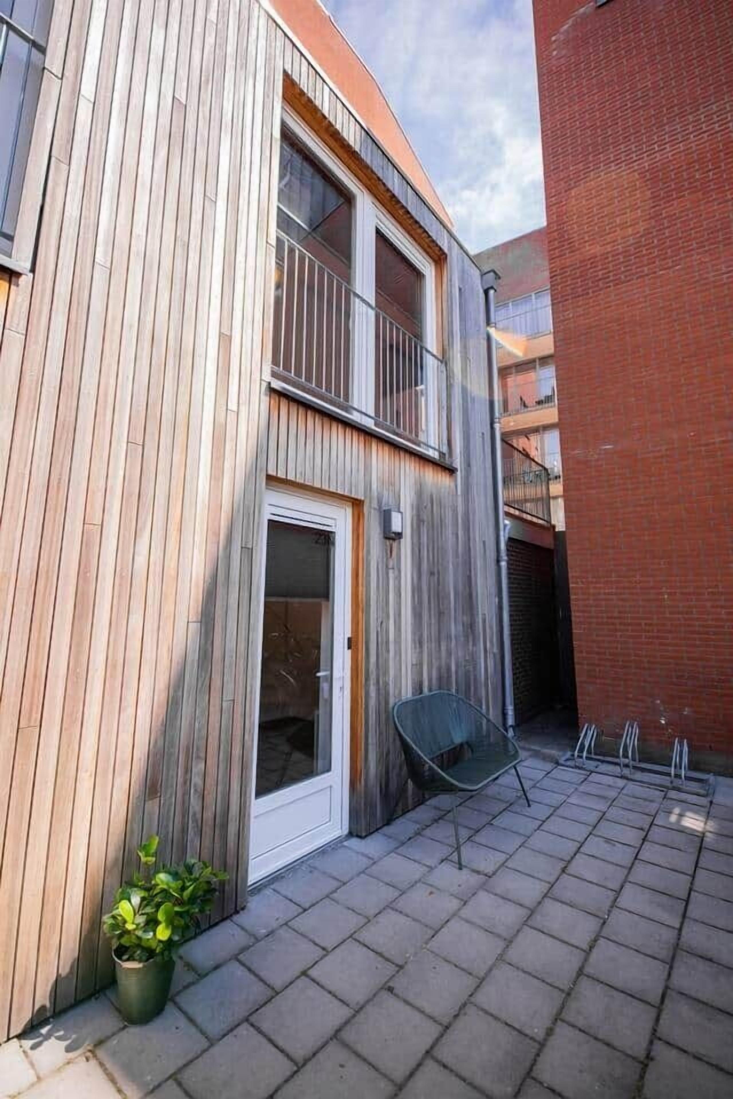 Un bâtiment moderne avec une façade en bois, une entrée avec une porte en verre et un petit espace extérieur aménagé avec une chaise et un pot de fleurs.