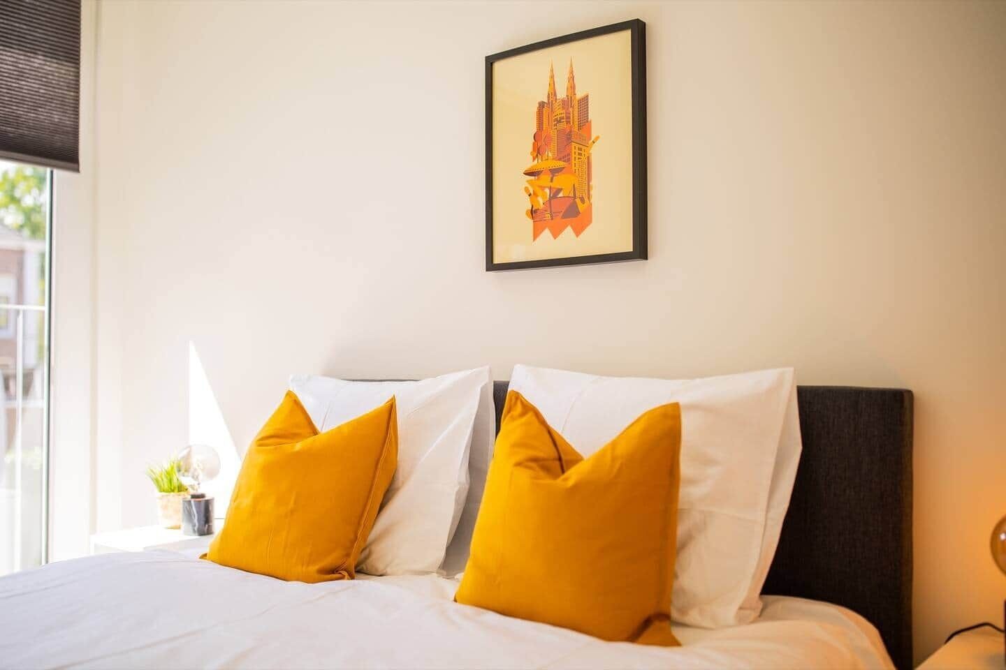 Ein Hotelzimmer mit Bett, weißer Bettwäsche, zwei gelben Kissen, einem gerahmten Kunstwerk an der Wand und einem Fenster mit Jalousien.