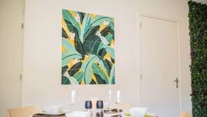Un comedor con una mesa redonda dispuesta para dos personas, un cuadro con motivos tropicales arriba y una pared cubierta de hojas verdes a la derecha.