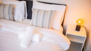 Un lit bien fait, avec du linge de lit blanc et des oreillers à rayures, une lampe de chevet et une table de chevet blanche.