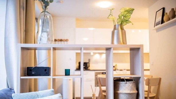 Un salon moderne avec un canapé gris, une console blanche agrémentée de plantes décoratives et, au fond, un espace cuisine.