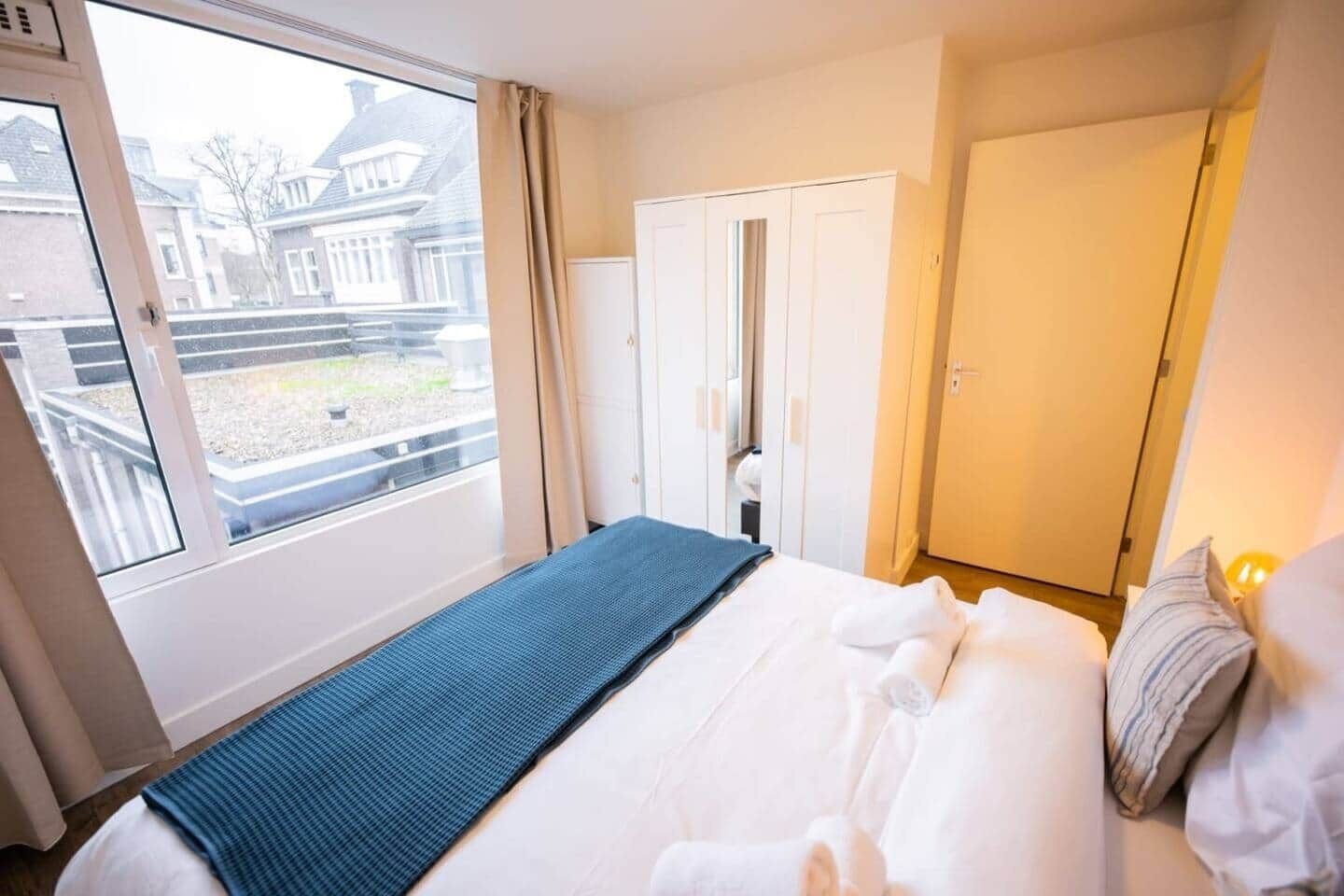 A hotel room with a large bed, a window with a view of buildings, a wardrobe, and a bedside table.
