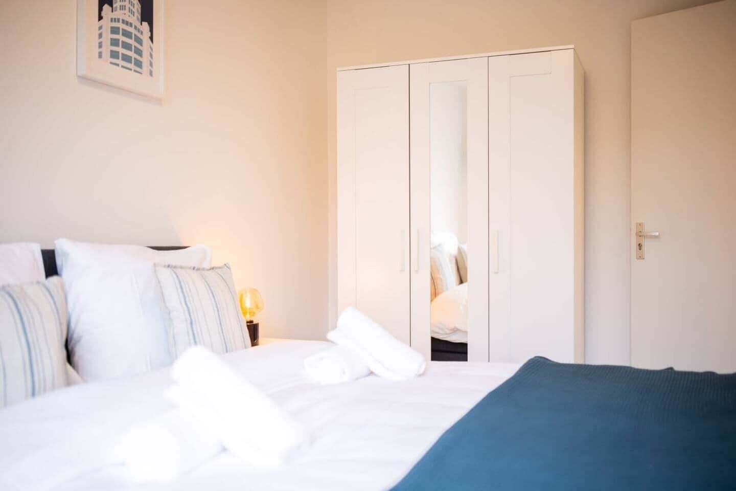 A hotel room with a bed, pillows, a bedside table with a lamp, and a wardrobe.