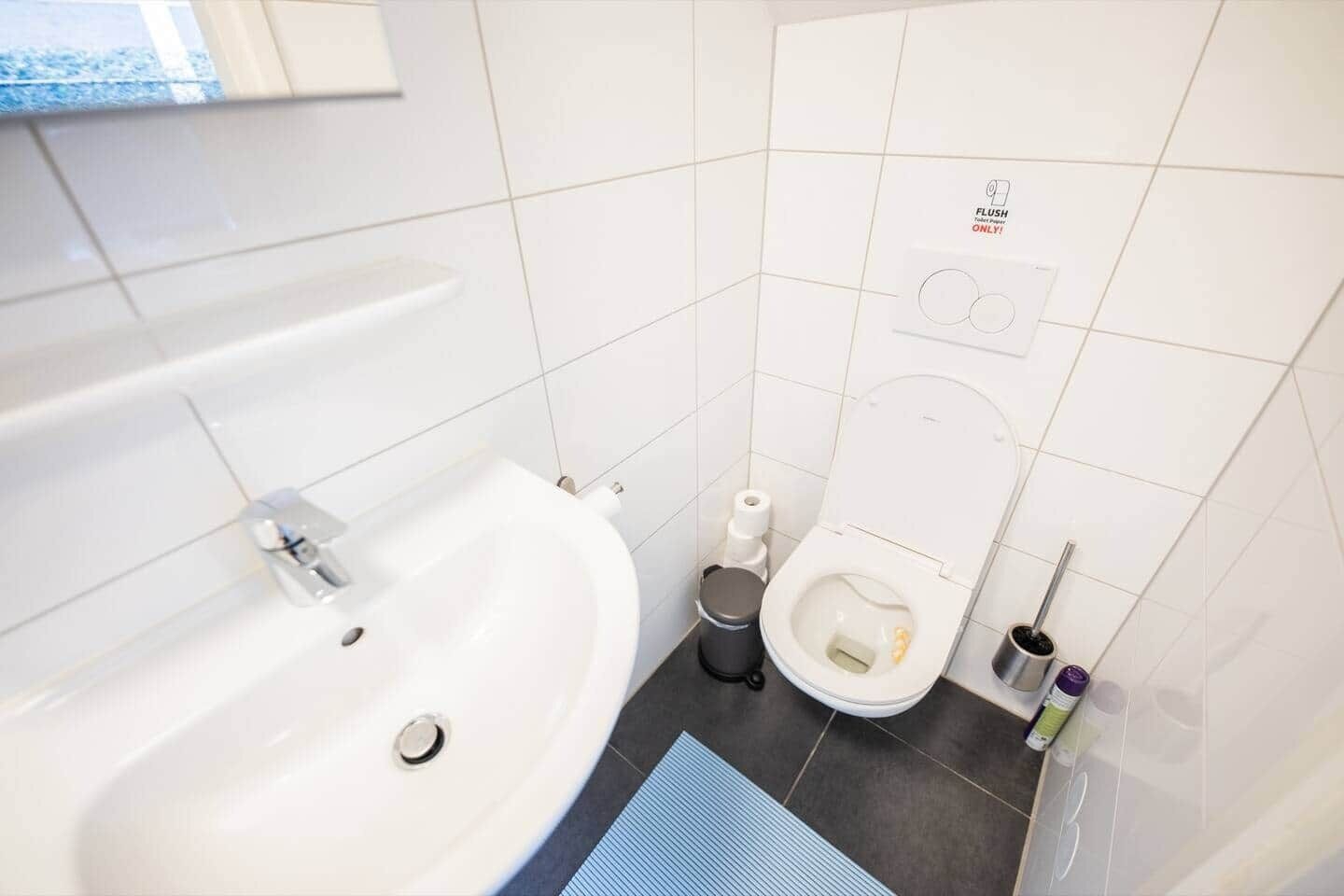 Een moderne badkamer met een wit wastafel, toilet en bidet. Er staat een prullenbak en een toiletborstel op de vloer.