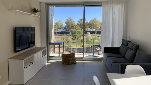 Ein modernes Wohnzimmer mit Flachbildfernseher, grauem Sofa, Esstisch und Balkon mit Blick auf einen Spielplatz und Bäume.