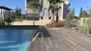 Eine Holzterrasse führt zu einem Schwimmbecken mit einem Metallgeländer, umgeben von einer angelegten Grünfläche mit Bäumen und einem Gebäude im Hintergrund.