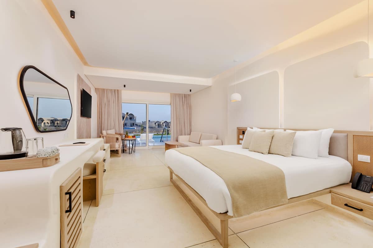 A modern hotel room with a large bed, a desk, a chair, a small table, and a view of the city.