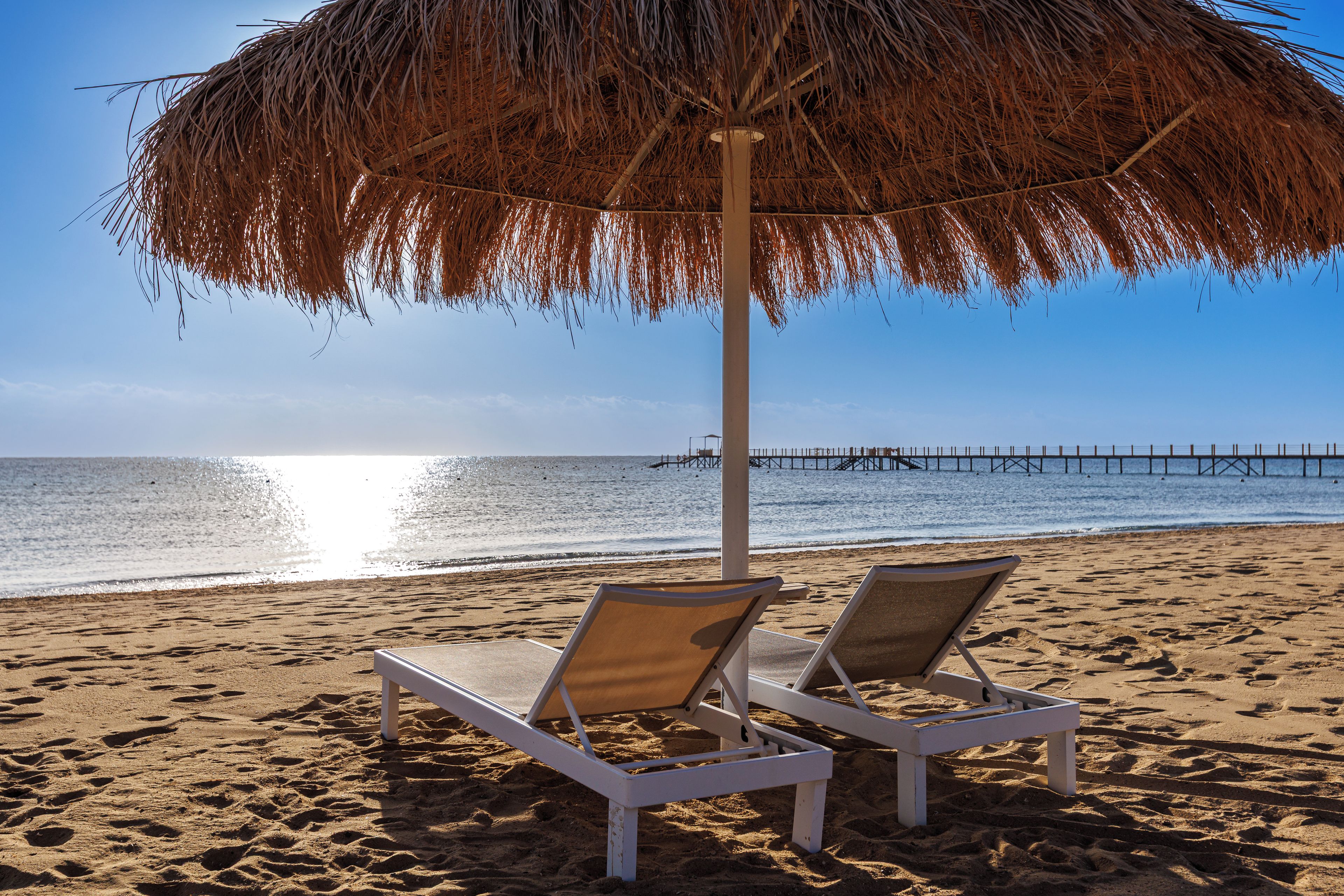 A beach with two lounge chairs under a thatched umbrella, a pier in the background, and clear skies.