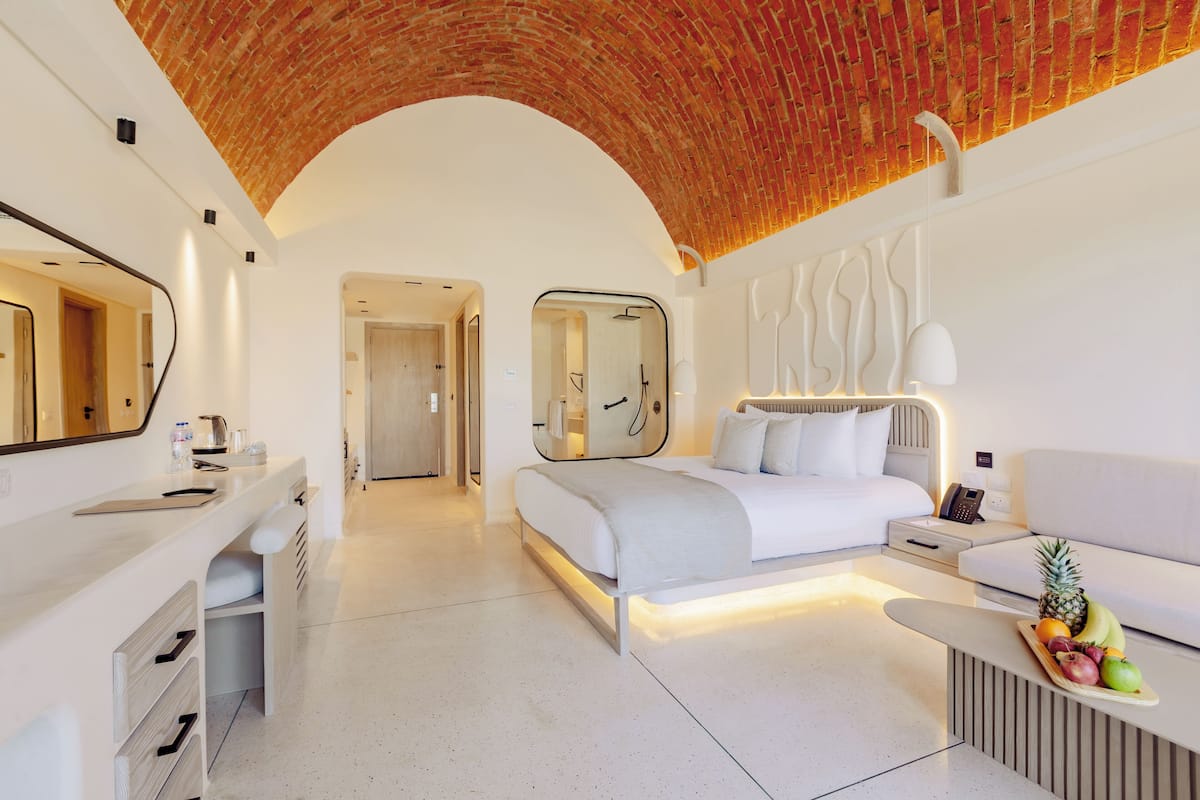 A modern hotel room with a brick ceiling, a large bed with white linens, a bedside table with a fruit tray, a white sofa, and a bathroom visible through an archway.