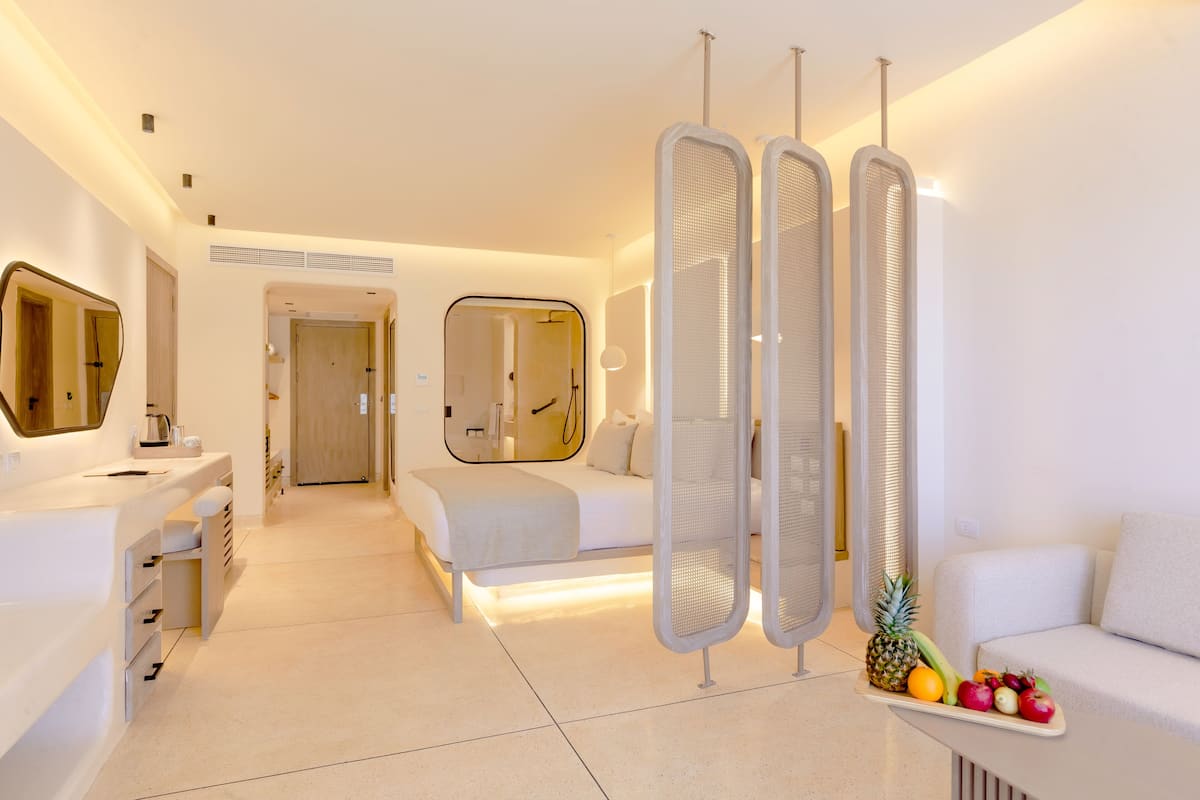 A modern hotel room with a large bed, a sitting area, a bathroom with a mirror, and a fruit tray on the side.
