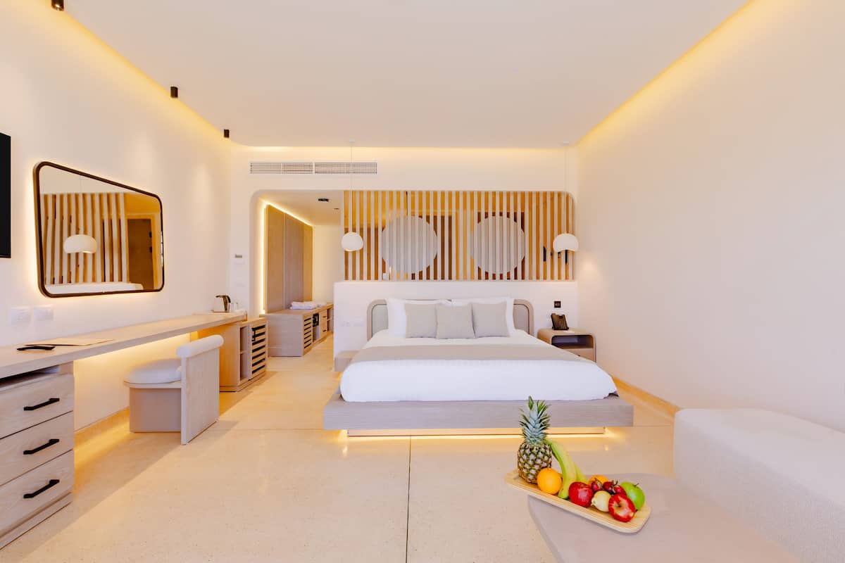 A modern hotel room with a large bed, a desk, and a vanity. There is a fruit tray on the bed and a TV mounted on the wall.