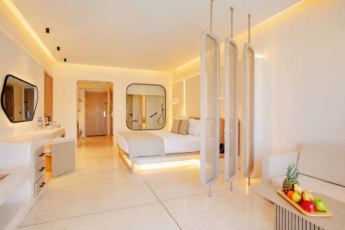 A modern hotel room with a bed, a sitting area with a tray of fruit, a mirror, and a door leading to another room.