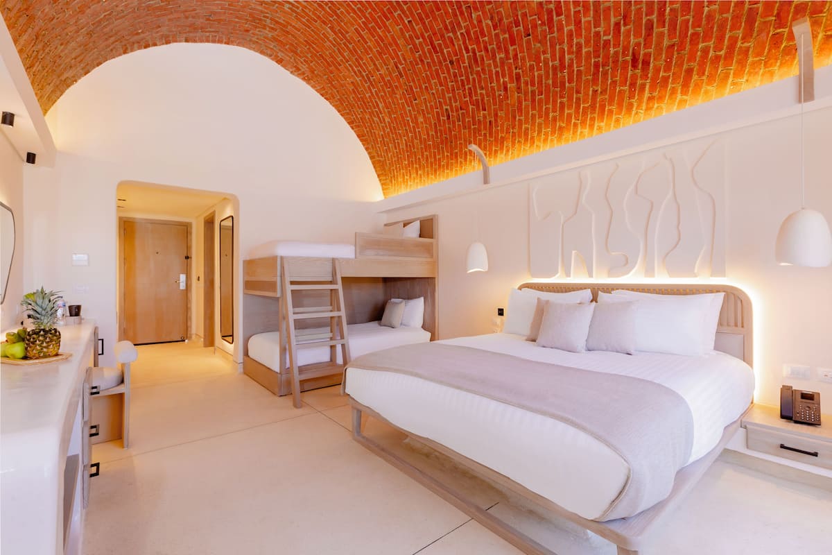 A modern hotel room with a large bed, a bunk bed, a desk with a chair, and a brick ceiling.