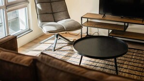 Una sala de estar con un sofá de cuero, una mesa de centro negra, un sillón gris, un mueble bajo para la televisión de madera y una lámpara de pie.