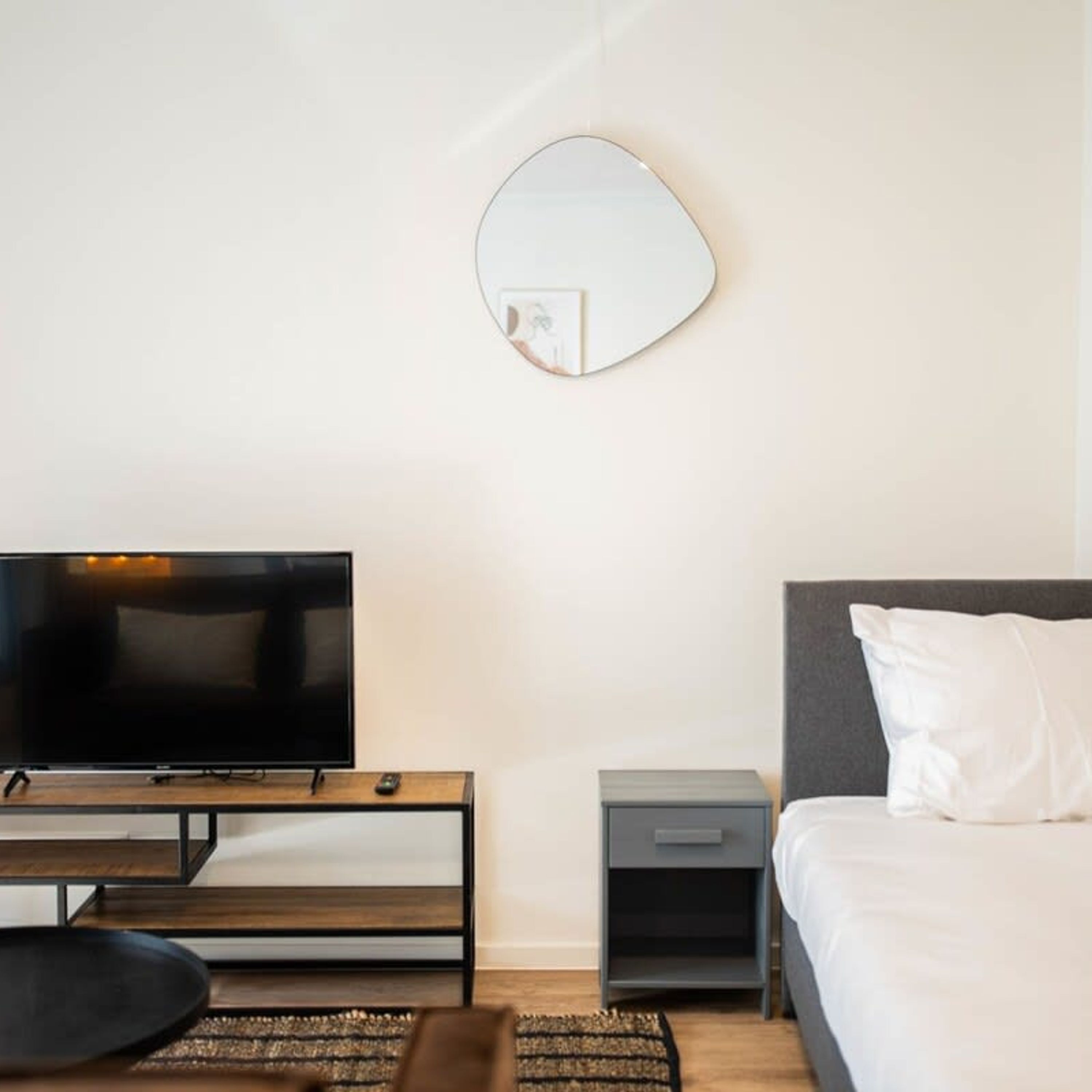 A modern hotel room with a flat-screen TV, a bedside table, a small drawer, a round mirror, a bed with a white pillow, and a dark-colored headboard.
